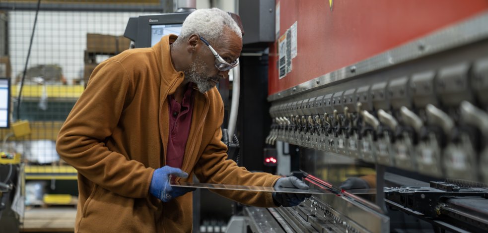 A worker wearing safety glasses and gloves operating a CNC press brake to bend a sheet metal part.