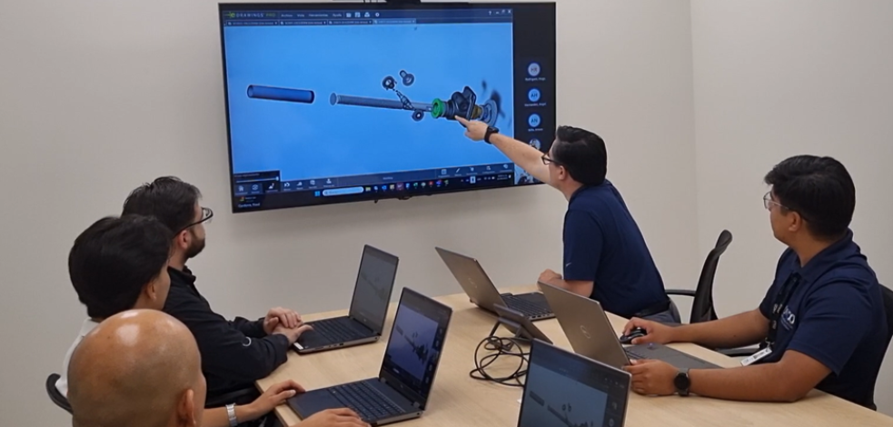 A team of engineers reviewing a 3D CAD model of a mechanical assembly on a wall-mounted monitor while collaborating on laptops around a conference table.