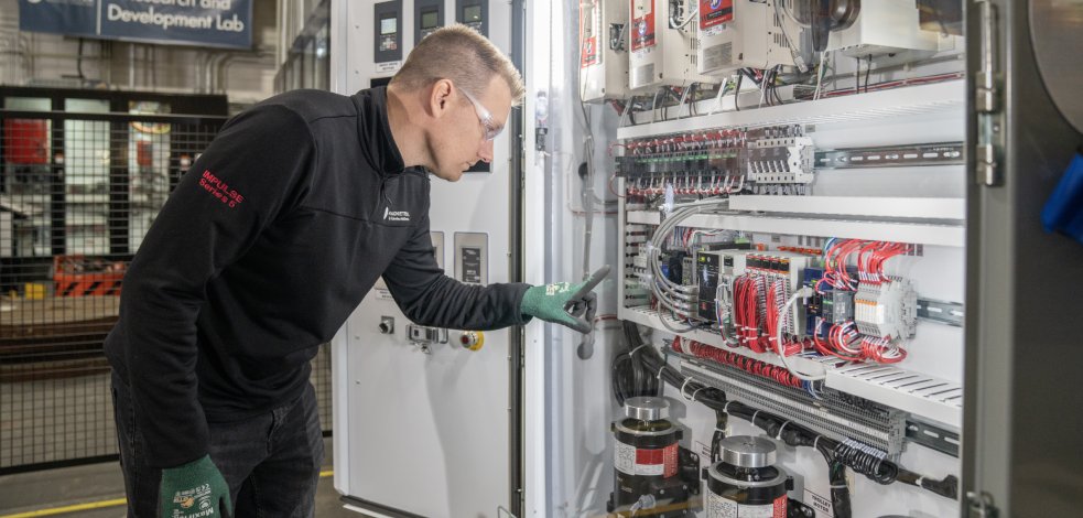 A technician in safety glasses inspecting the wiring and control components inside an open industrial control panel.